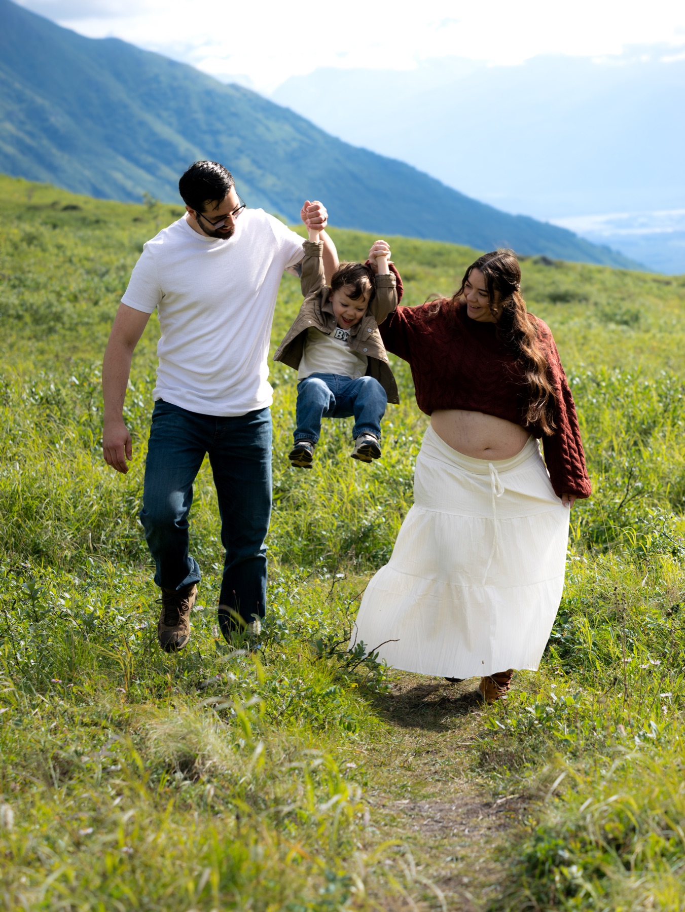Parents swinging their young child between them on a green mountain trail at Hatcher Pass, Alaska